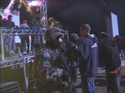 Maskierter Soldat mit Maschinengewehr, im Hintergrund weitere Soldaten und Raver auf und vor einer Konzertbühne
